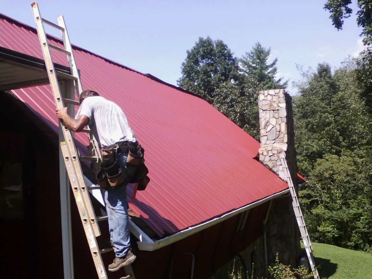 Roofer on a ladder beside a red metal roof and stone chimney — Soffit & Fascia Repair by Roofing Brothers in Isleton